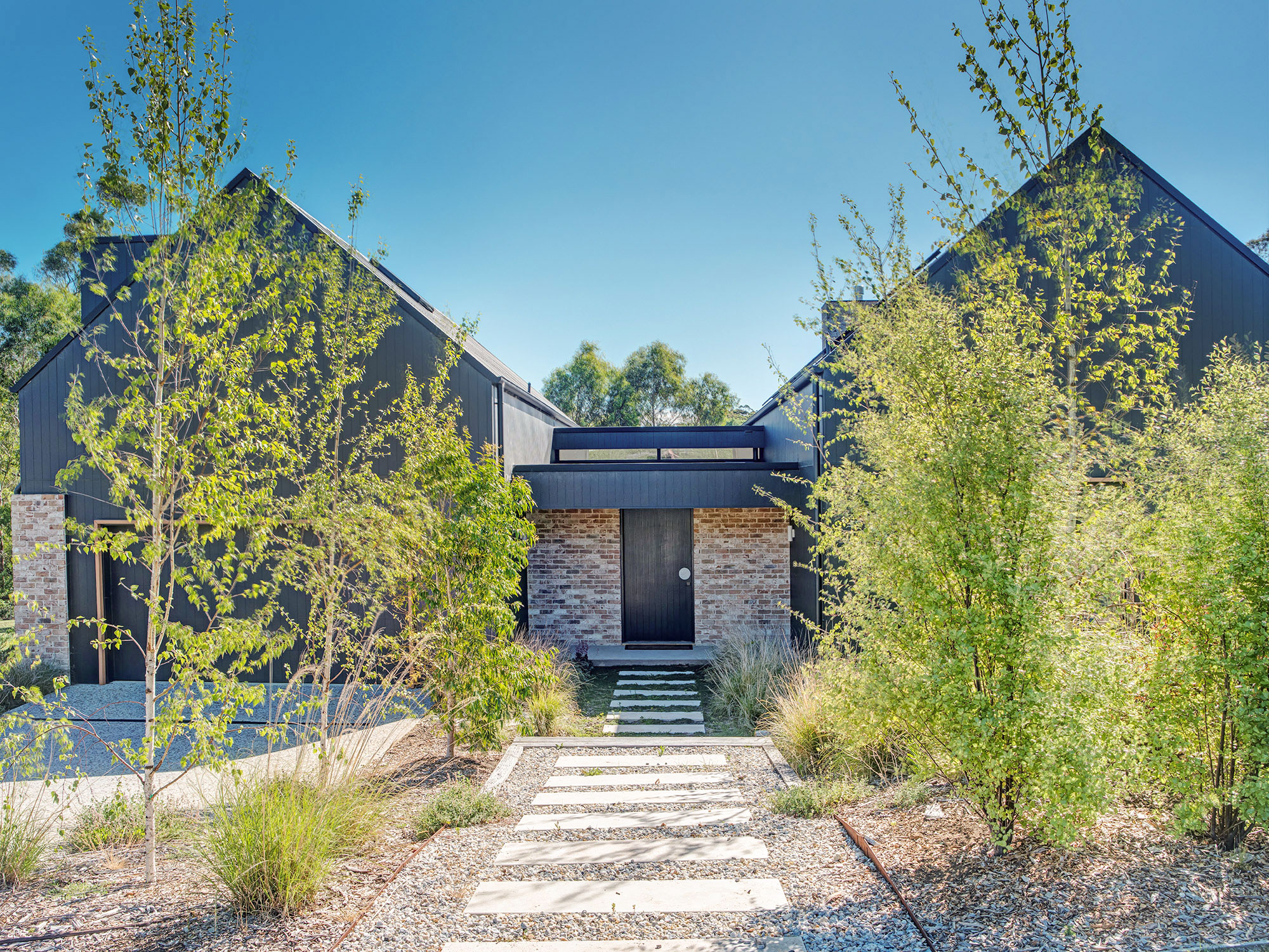 Modern pavilion home with recycled brick accents and large windows, surrounded by perennial gardens and trees under clear blue sky.