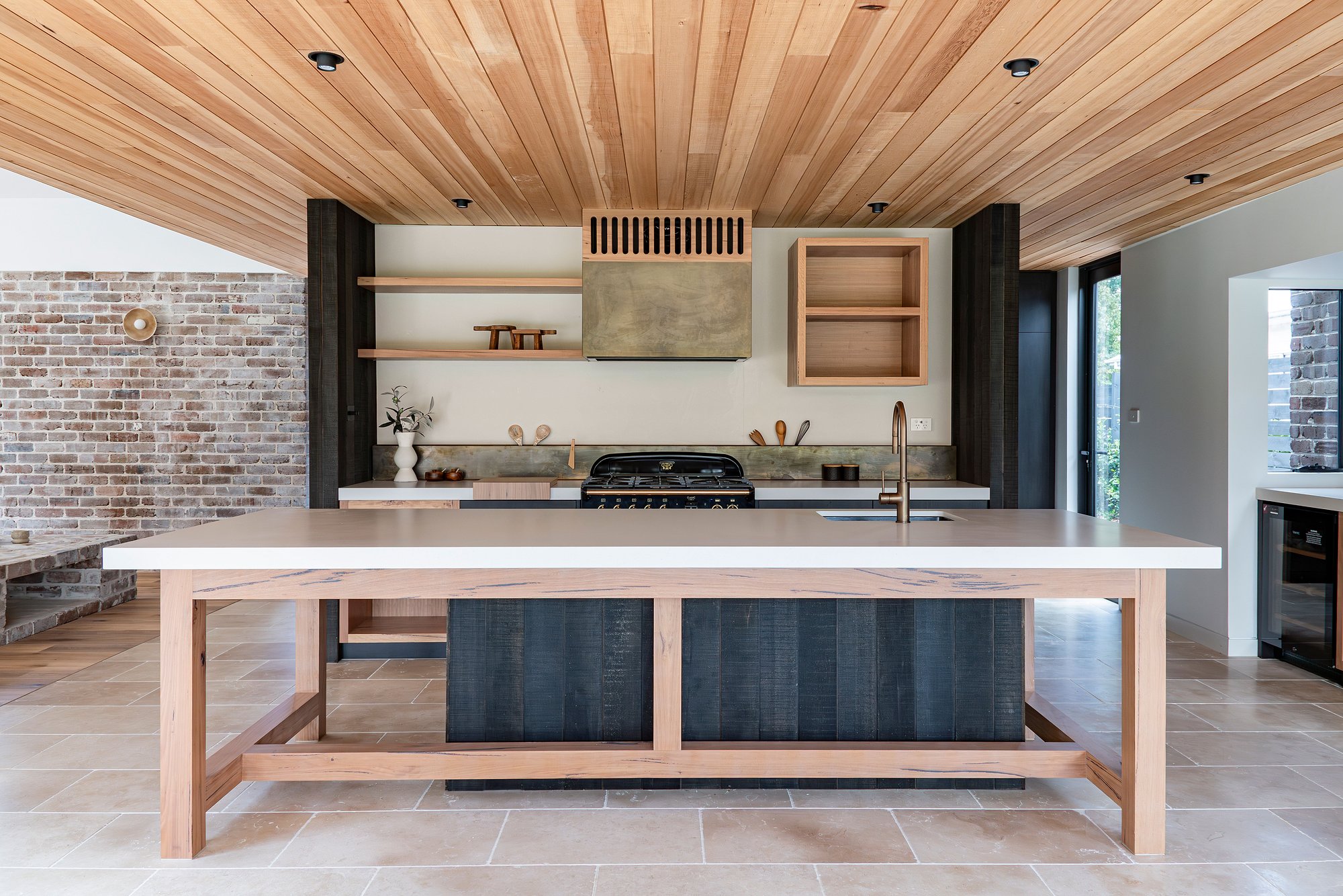 Modern kitchen with timber ceiling, exposed brick wall, stone benchtops and custom timber island featuring integrated seating and storage.