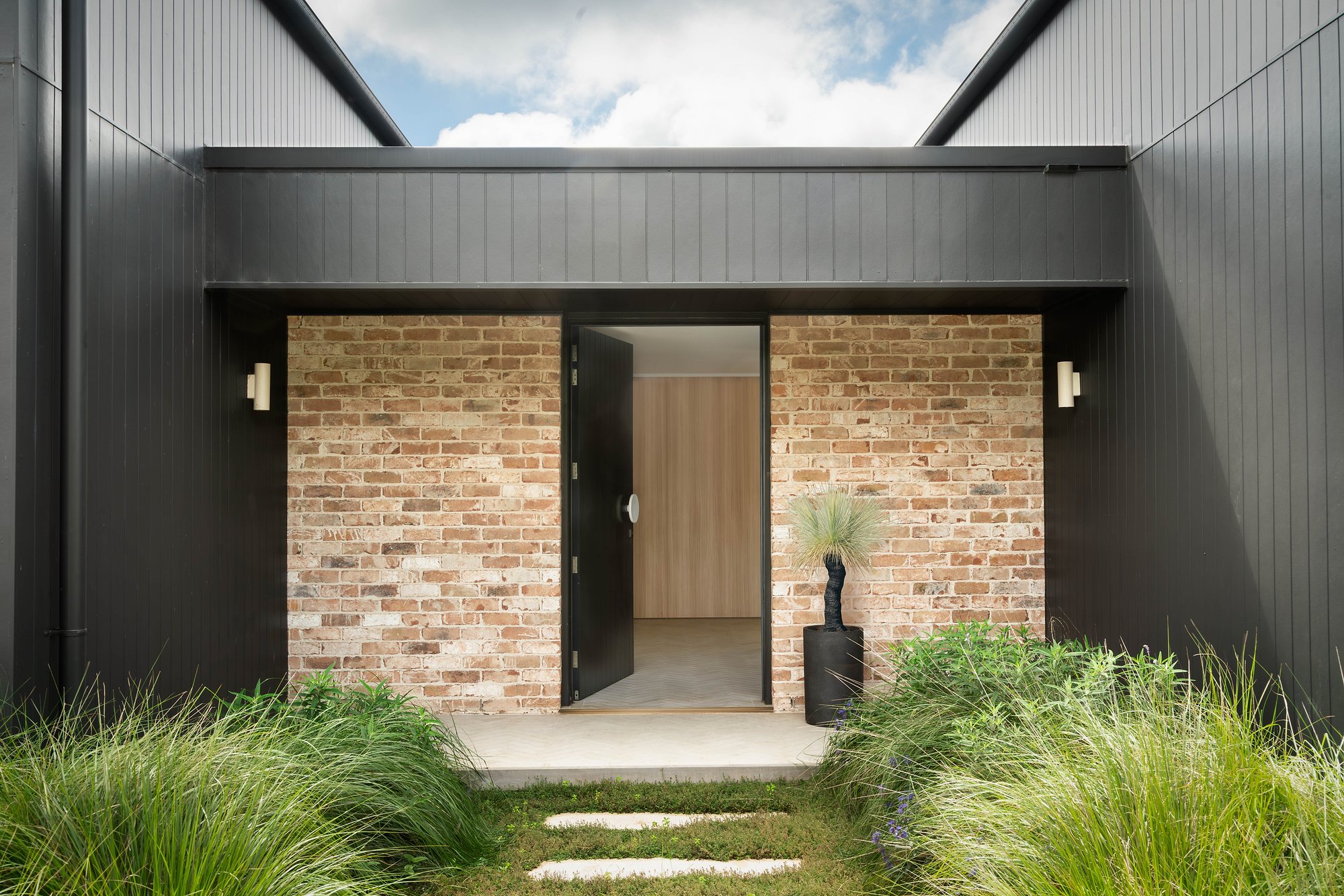 Recycled brick entry pavilion with charcoal steel cladding and brass hardware, flanked by ornamental grasses.
