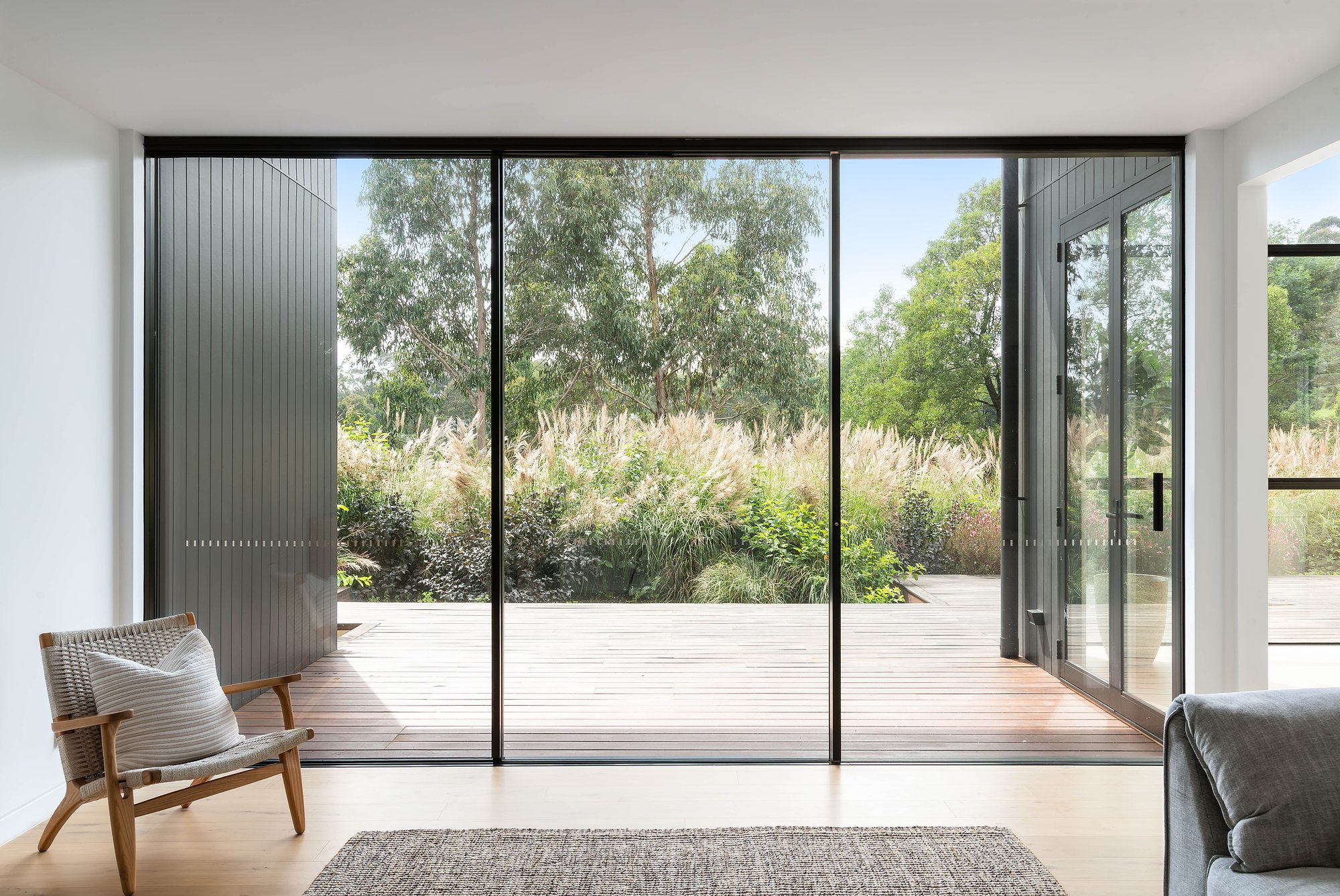 Floor-to-ceiling glazing frames native grassland beyond timber decking, with fluted glass privacy screen and mid-century armchair in foreground.