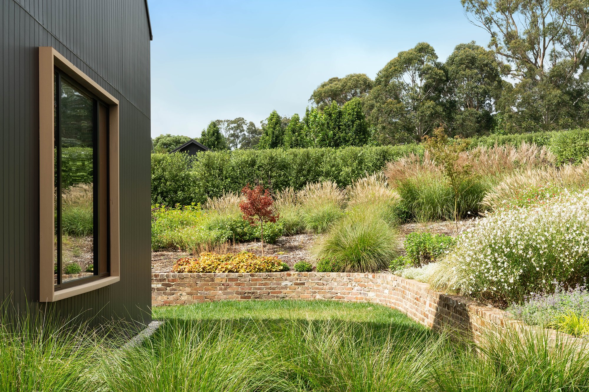 Modern pavilion with charcoal cladding overlooks native grasses and recycled brick retaining walls in terraced garden.
