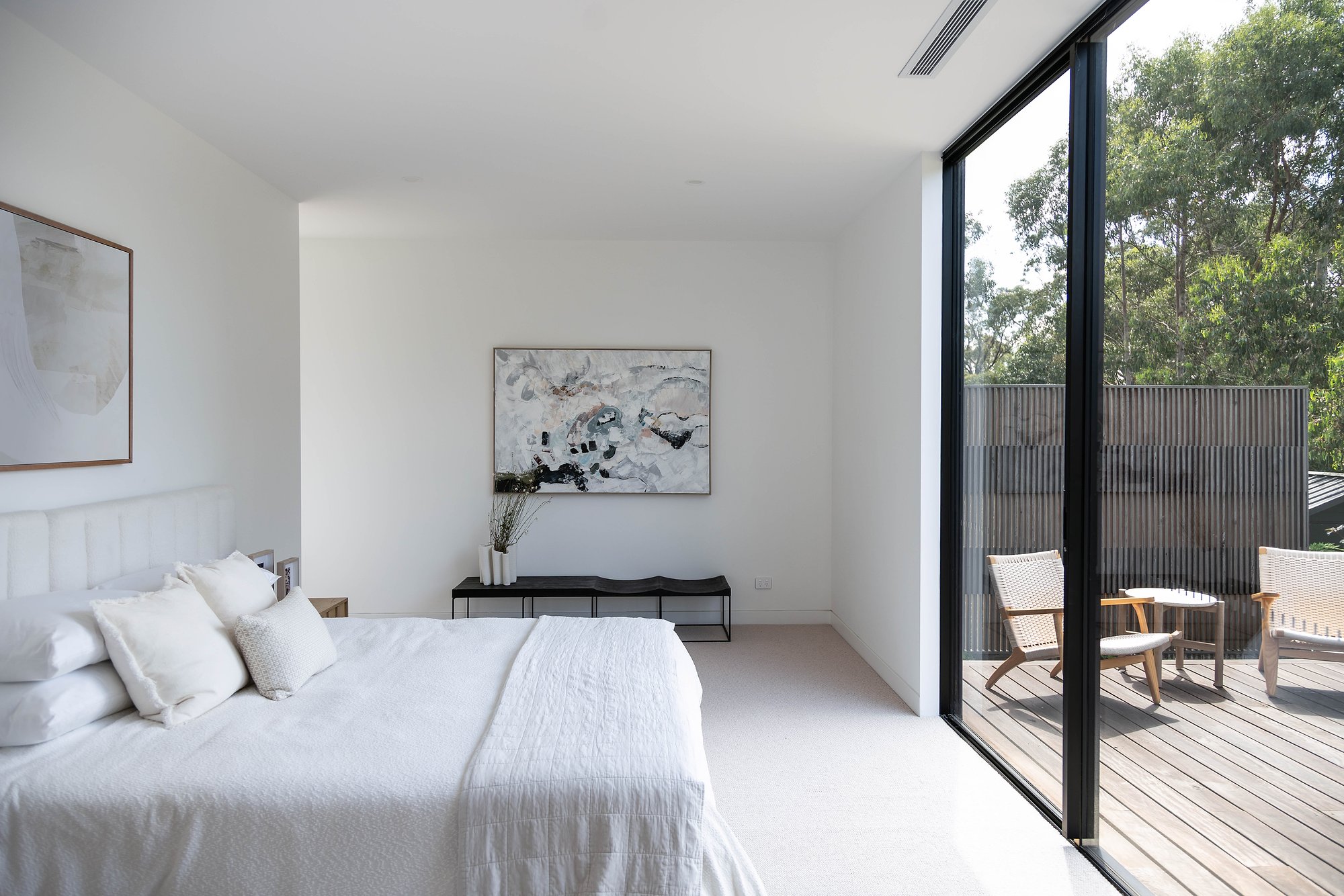 Bedroom with 100% wool carpet flooring opens to timber deck through floor-to-ceiling glazing, framing established trees beyond.