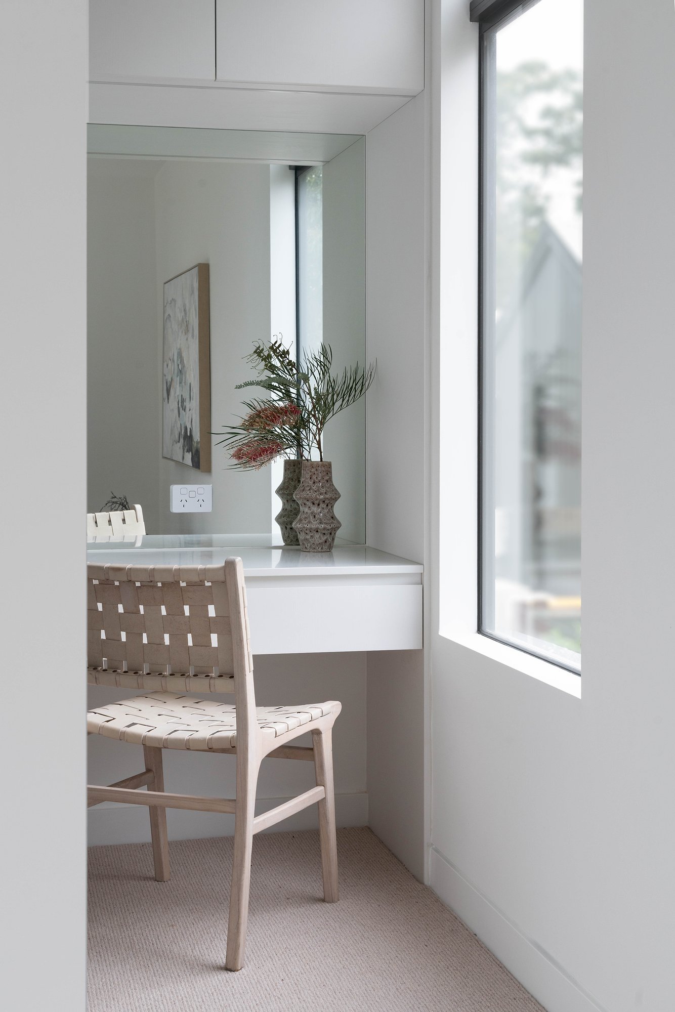 Built-in desk with woven timber chair beneath window, white joinery with ceramic vase and dried palm fronds.