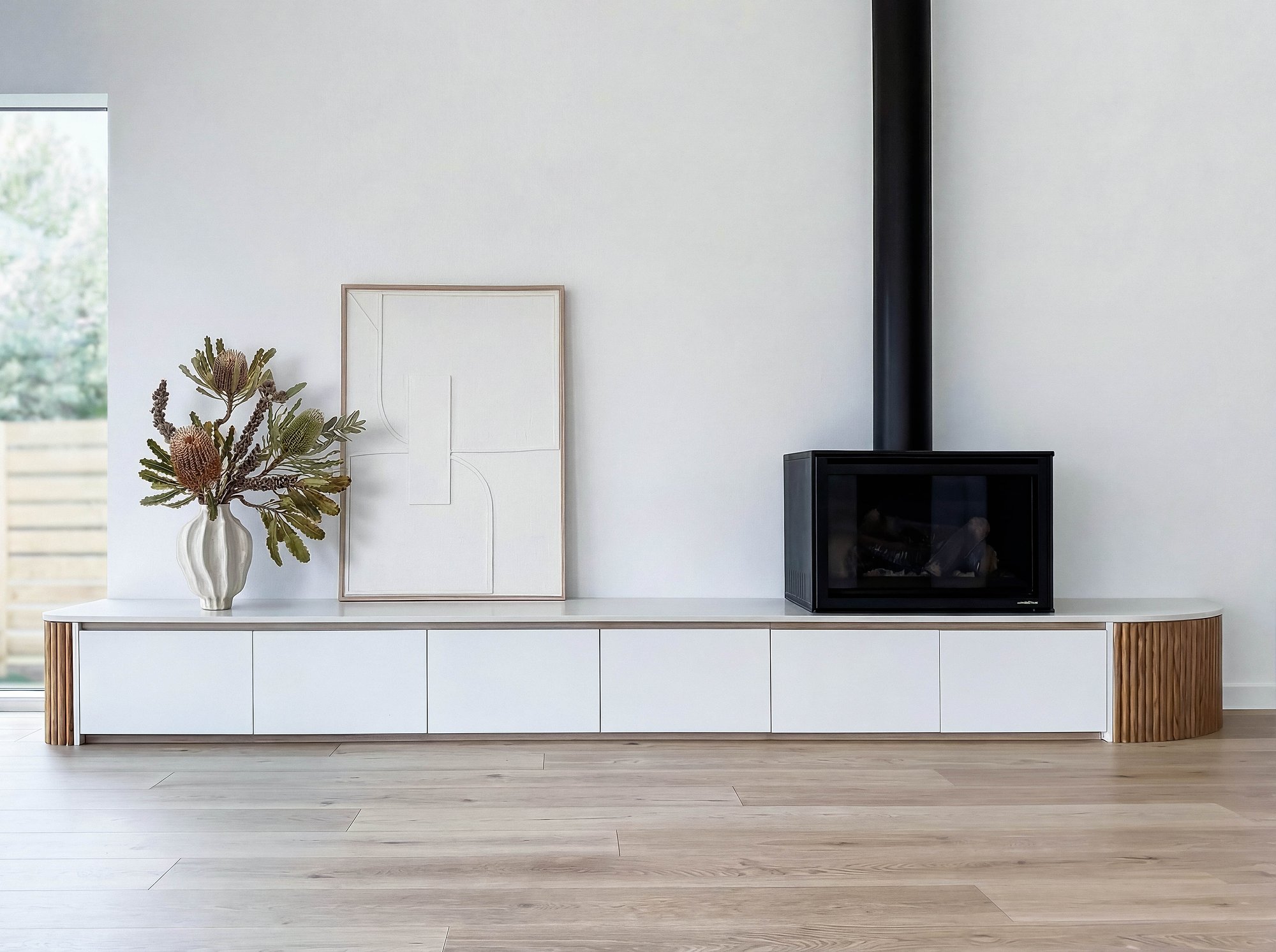 White media unit with fluted timber details beneath suspended black fireplace, styled with native foliage against pale walls.