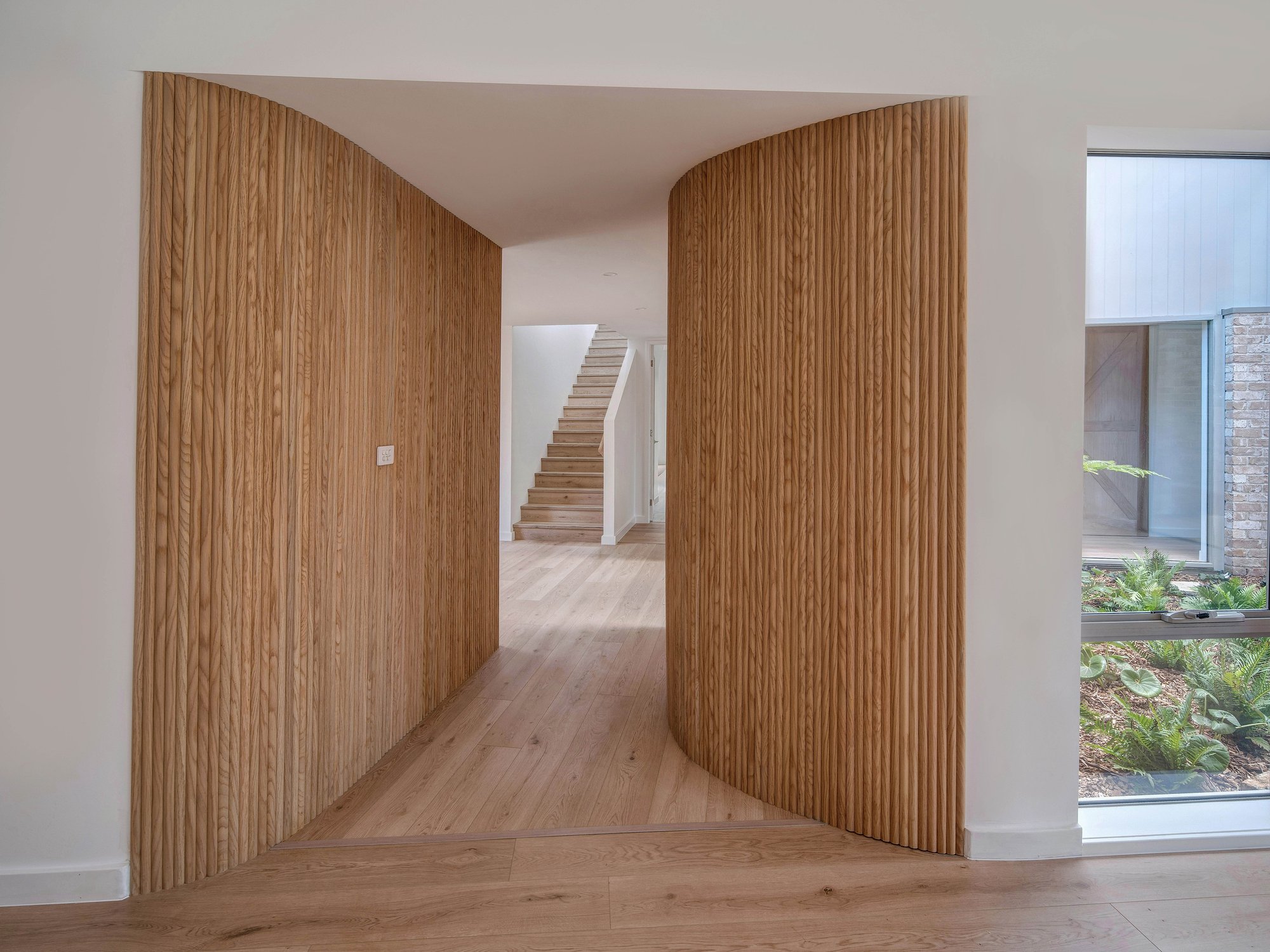 Curved timber-lined walls frame a hallway with white oak flooring, leading toward stairs and garden views.
