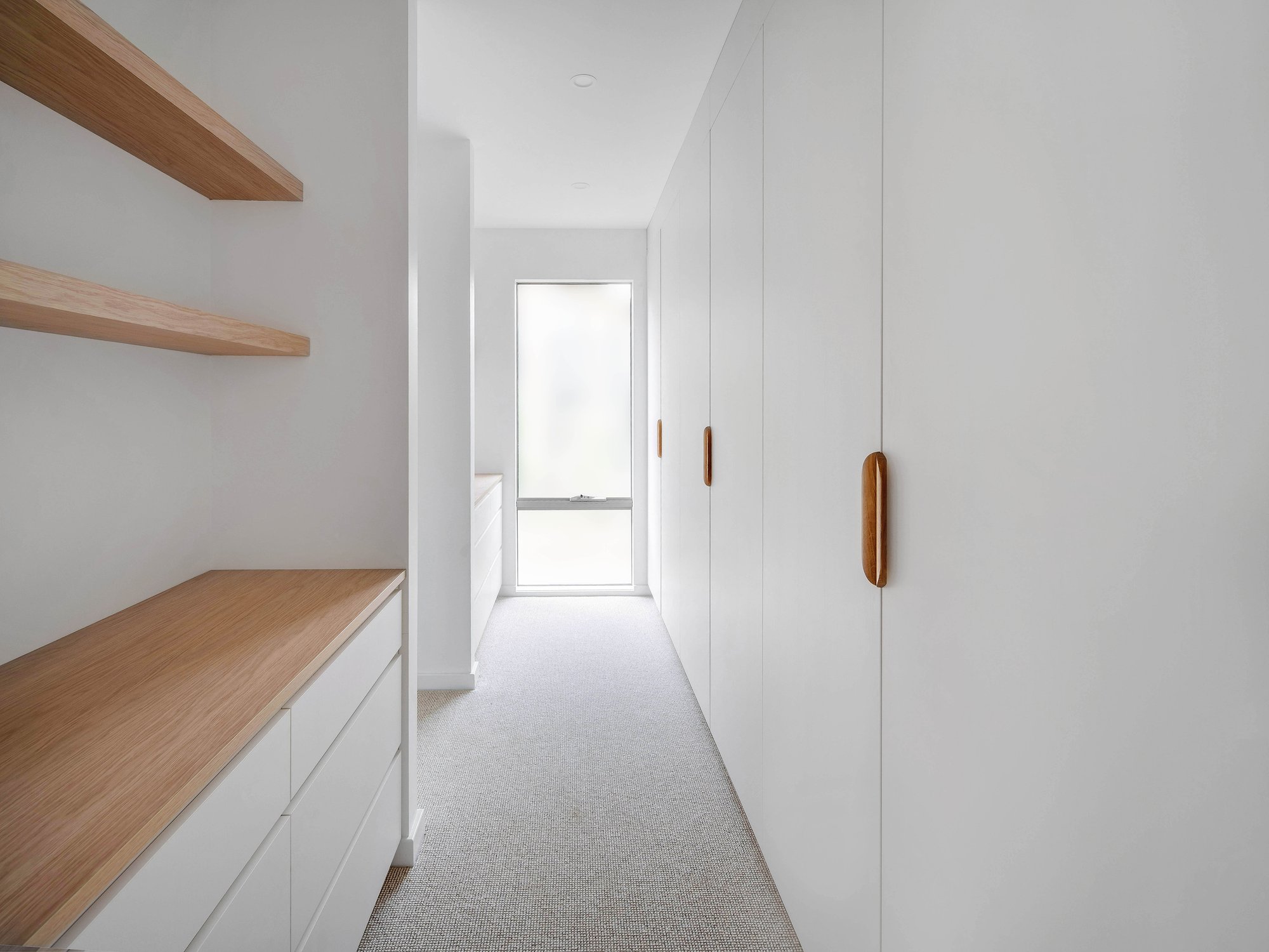 White hallway with wool carpet flooring, oak floating shelves, and integrated joinery leading to glazed rear doors.