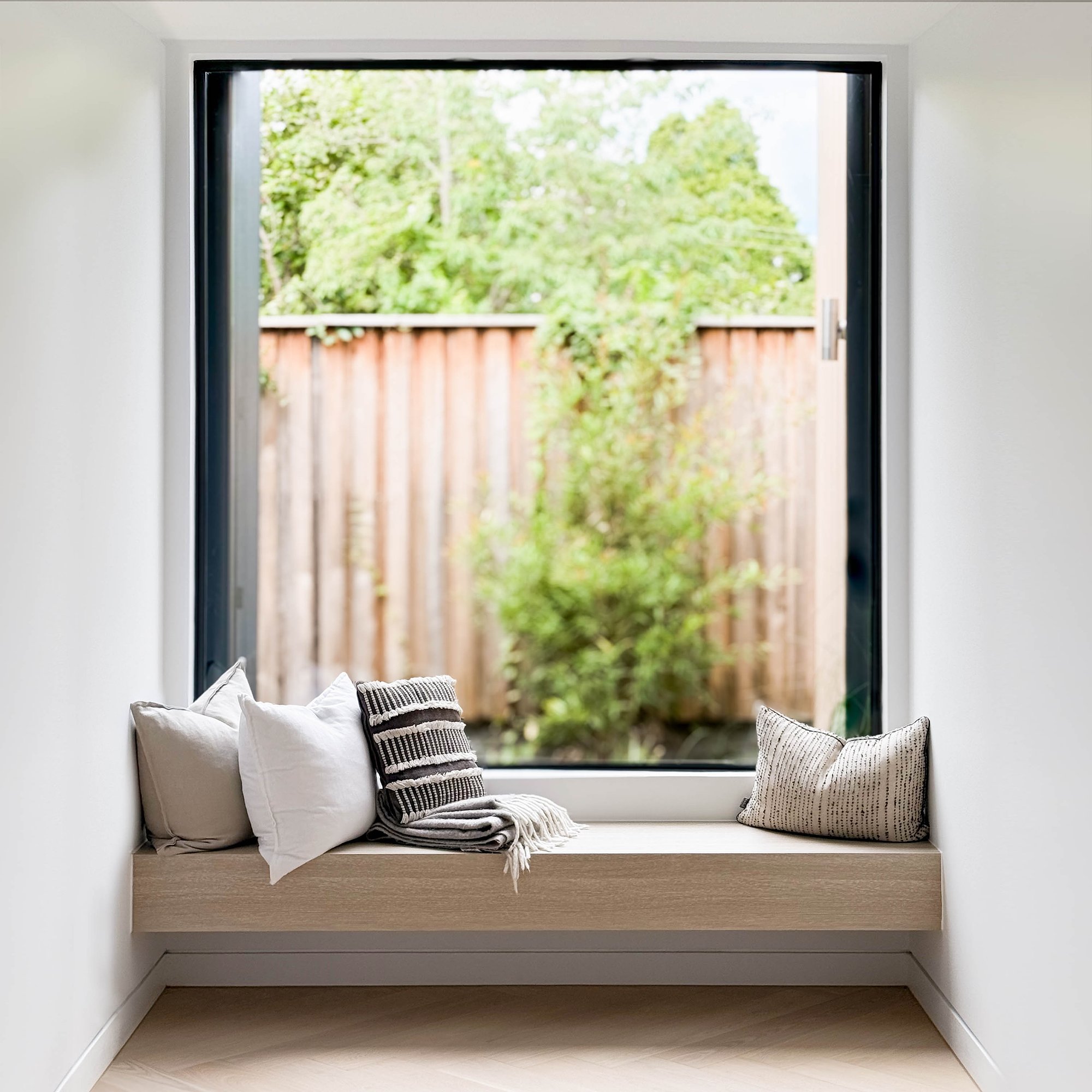 Built-in window seat with linen cushions and woven throw, overlooking timber-battened fence and established garden plantings.