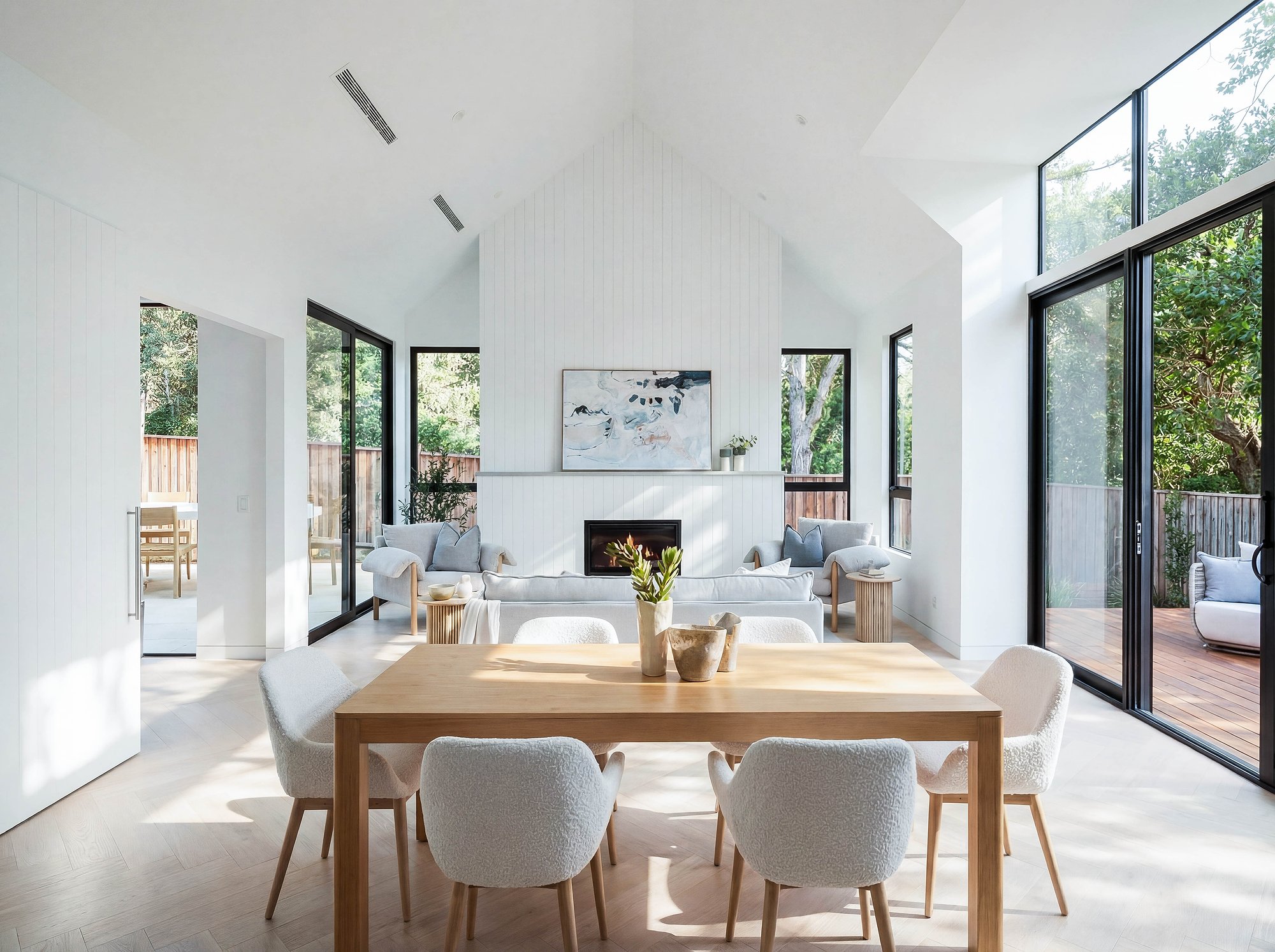 Open-plan living with timber dining table, concrete fireplace, and floor-to-ceiling glazing framing established trees.