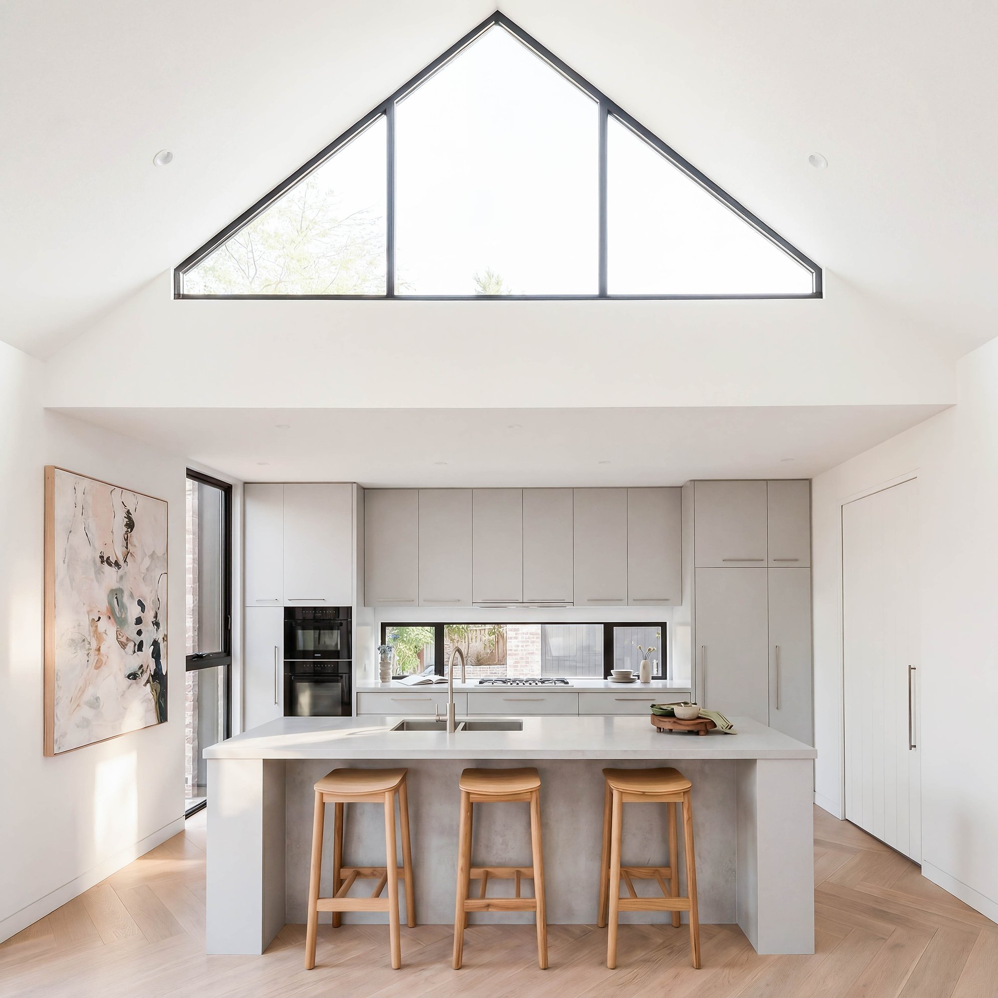 Kitchen island in reconstituted stone with timber bar stools beneath triangular clerestory window and integrated rangehood.