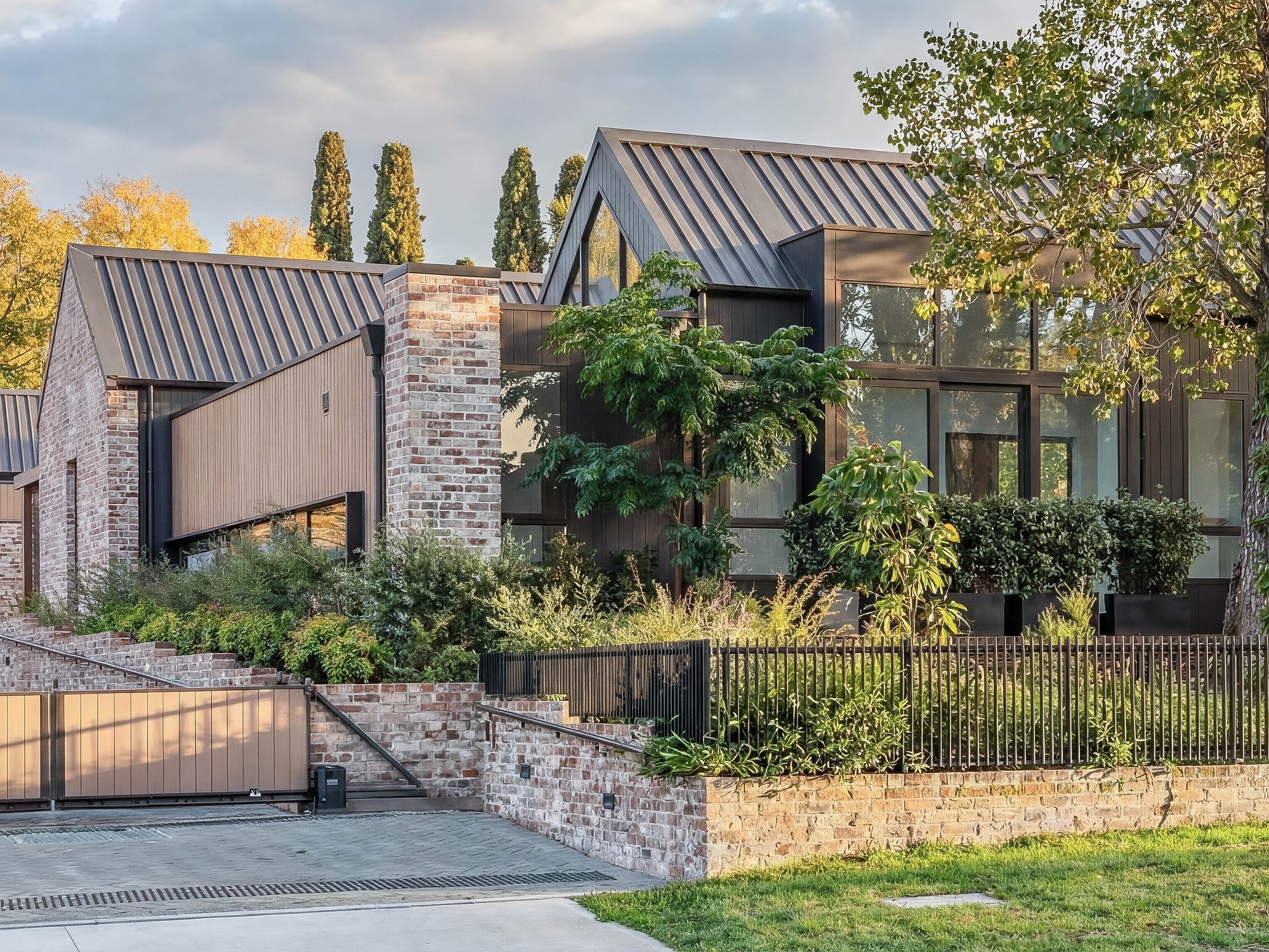Contemporary home with corrugated metal roofing, stone and timber cladding, and established garden plantings behind low stone wall.