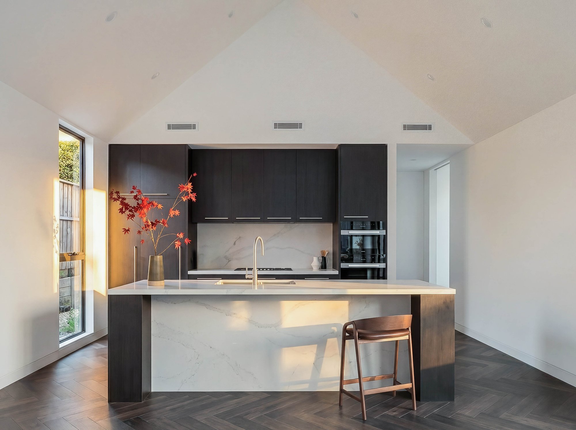 Kitchen island in honed stone with black cabinetry and timber barstool on polished concrete floors.
