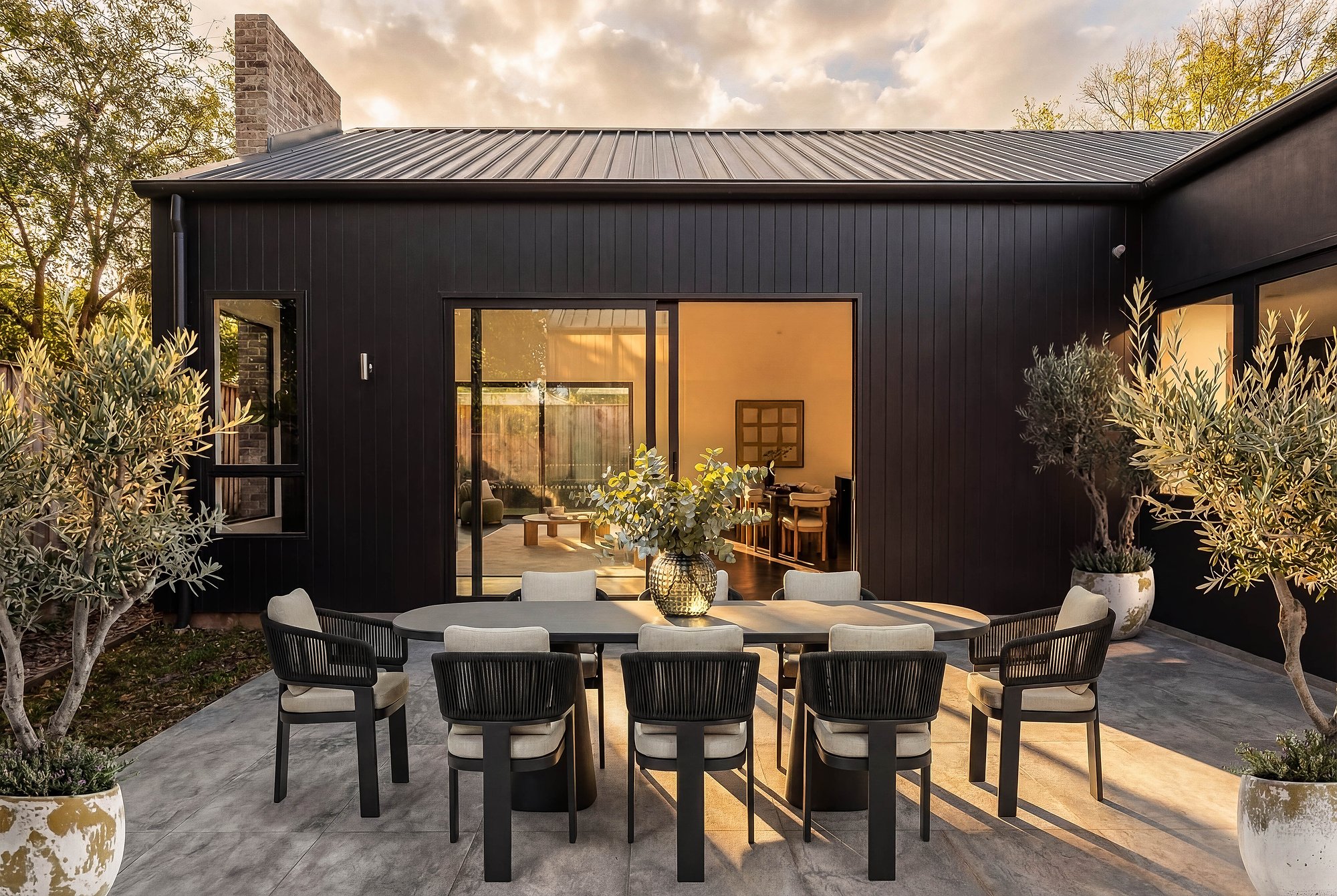 Black metal cladding pavilion with glazed doors opening to timber dining terrace, surrounded by established plantings at dusk.