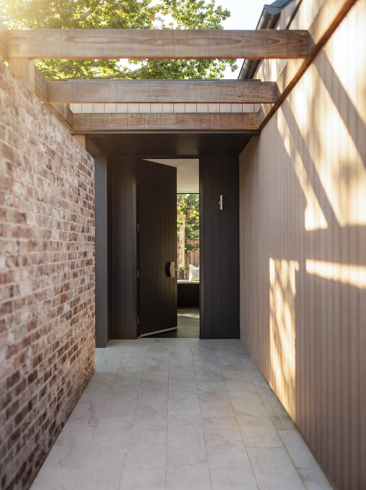 Recycled brick and timber cladding frame a dark steel entry door beneath an exposed concrete beam pergola.