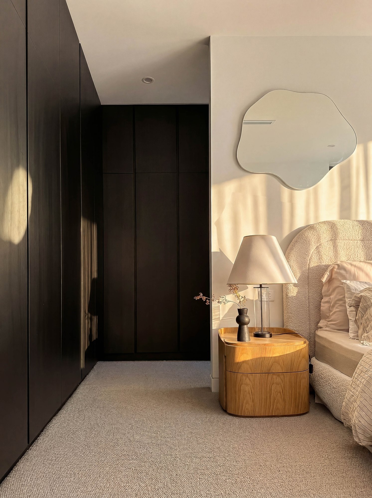 Bedroom with dark timber joinery, round American oak bedside table, and curved mirror catching afternoon light.
