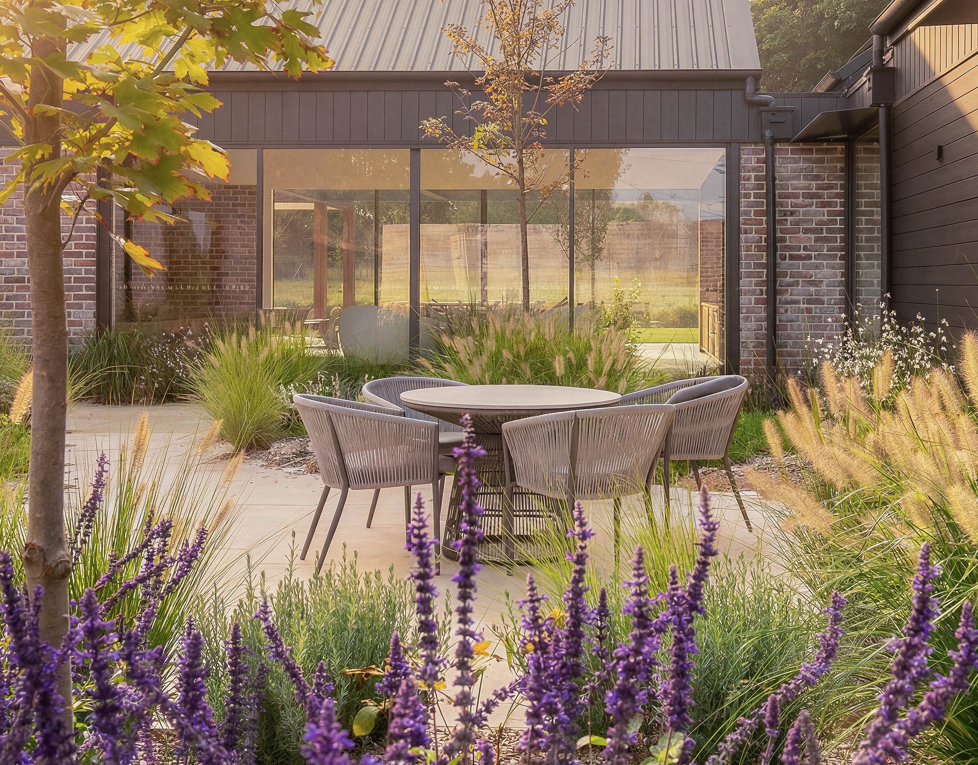 Contemporary pavilion with recycled brick walls and steel-framed glazing, surrounded by purple salvia and native grasses with timber outdoor dining setting.